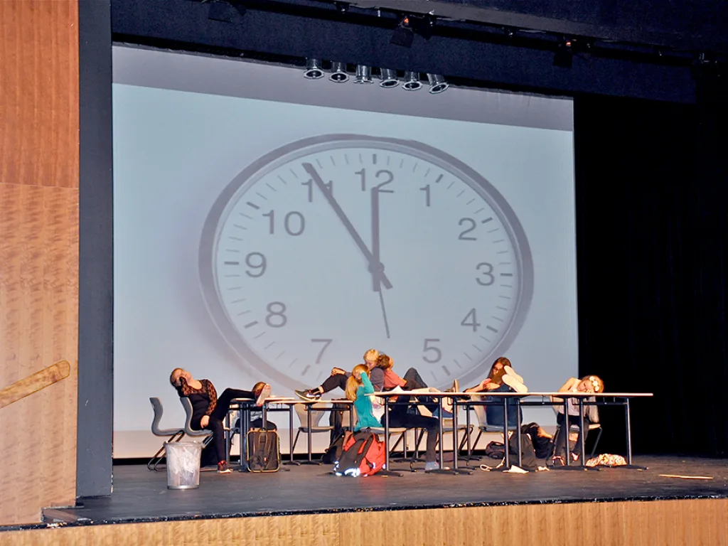 Darsteller sitzen an Tischen, im Hintergrund die projizierte Uhr.
